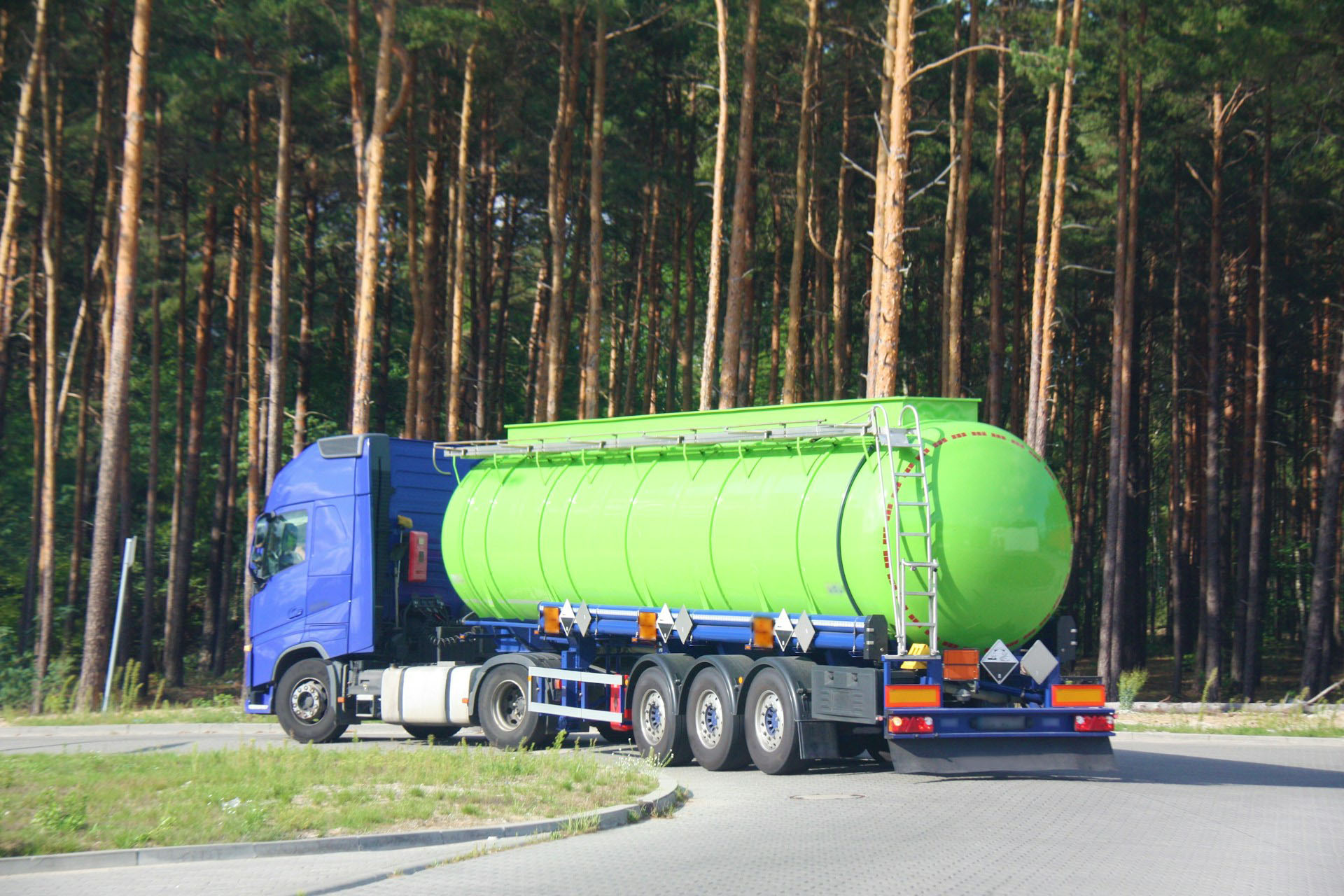 A blue and bright green tanker truck drives along a road that runs next to a forest.