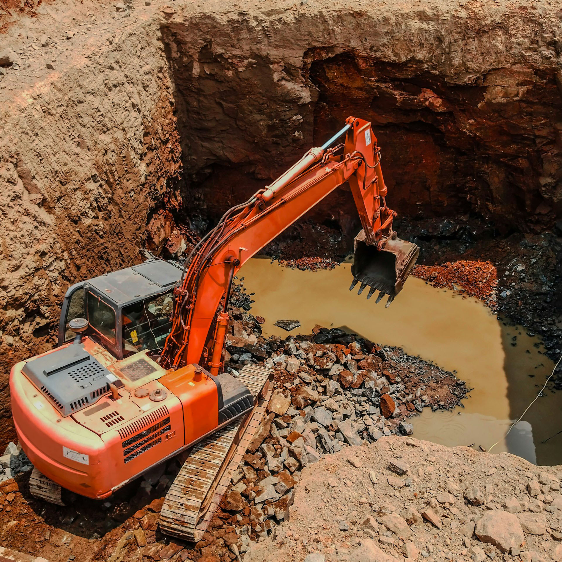 An orange excavator digging a hole with muddy water pooled at the bottom.