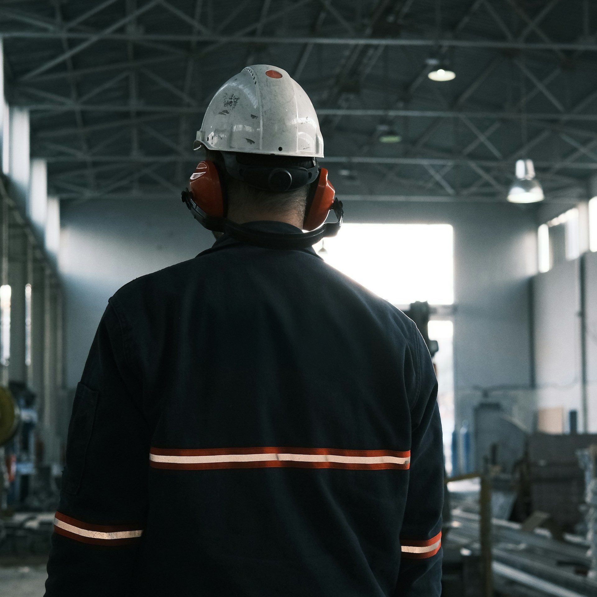 A construction worker wearing a dark blue jacket and white hard hat is facing away and looking out over an industrial facility.