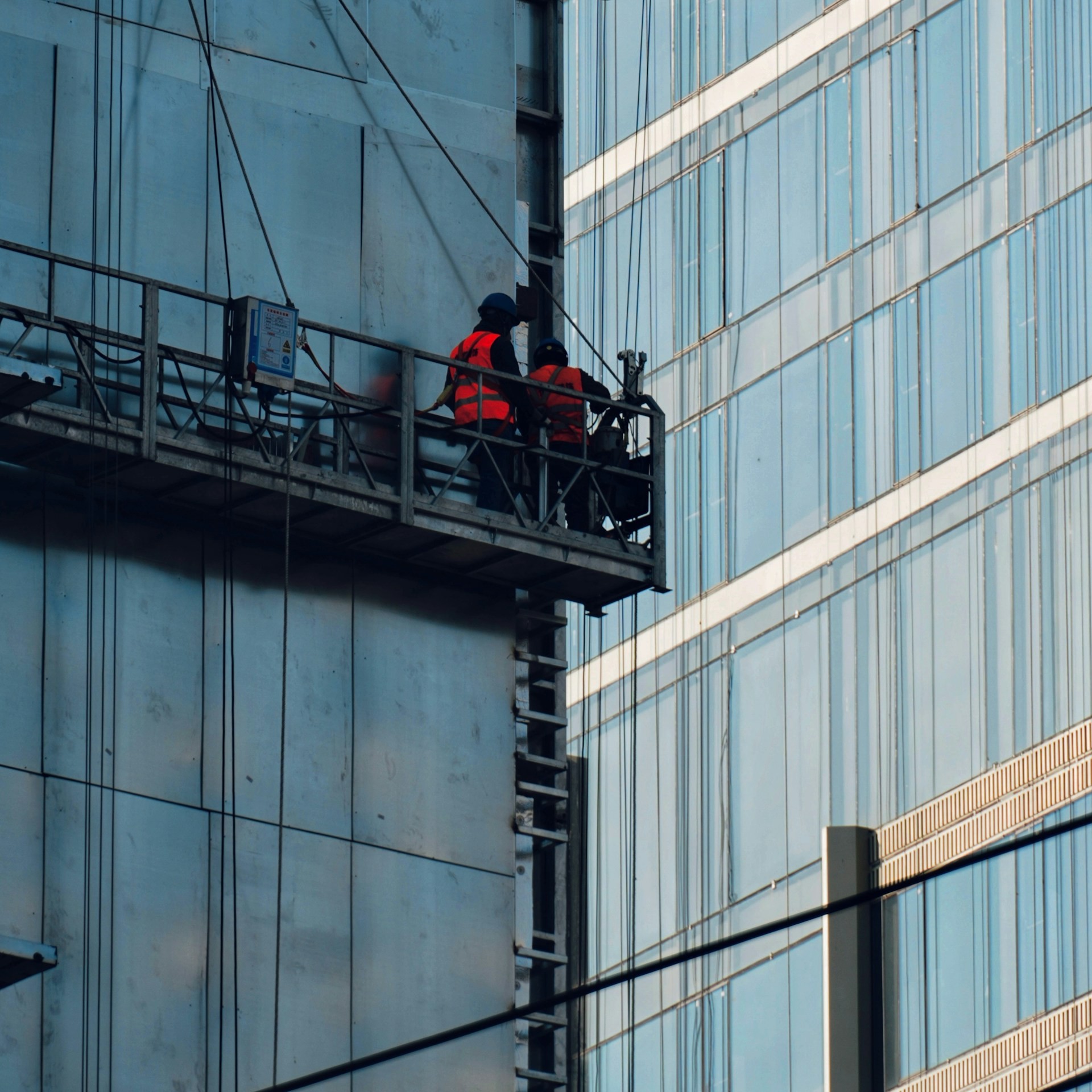 Two workers in bright orange vests stand on a work platform near the top of a tall glass building.